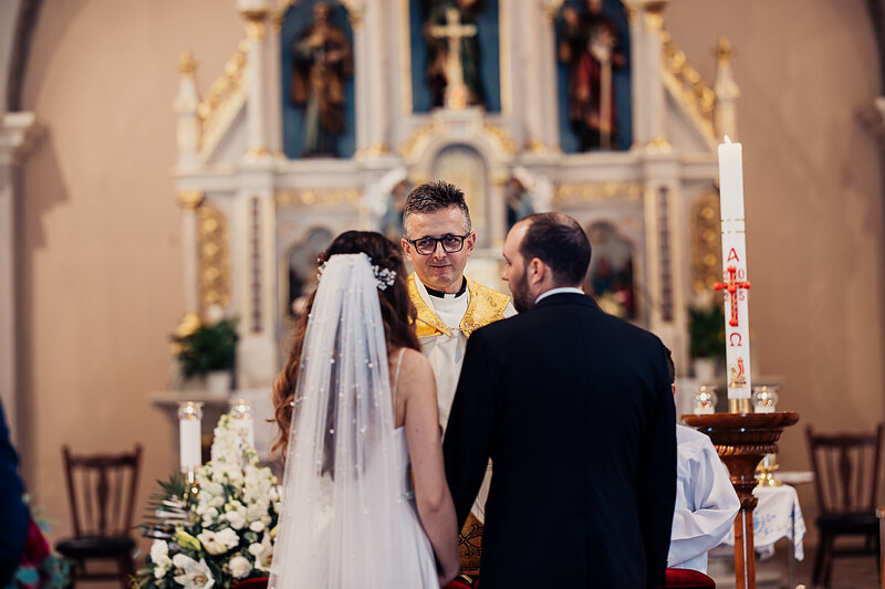 nevesta v závoji pri oltári, kňaz v zlatej ornáte, ženích a nevesta pred oltárom, sviečky v kostole, biele kvety na oltári, slávnostný obrad v chráme. Agropenzión Hubert, Ružindol.