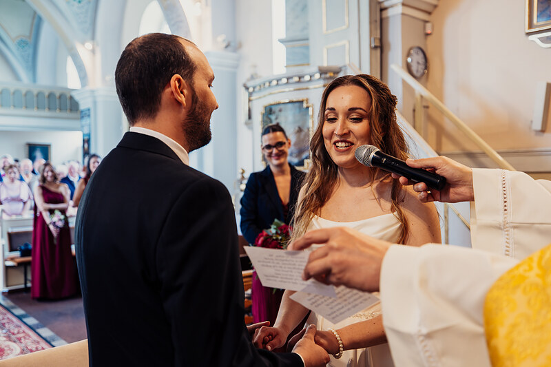 nevesta so závojom pri mikrofóne, ženích v čiernom obleku, kostolný obrad, kňaz v žltom ornáte, hostia v pozadí, prirodzené svetlo cez okná. Agropenzión Hubert, Ružindol.