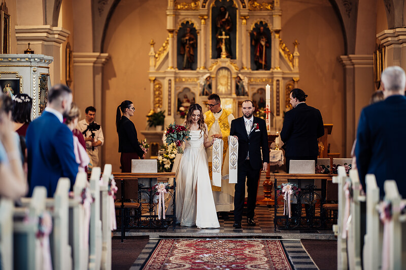 nevesta v bielych šatách, ženích v čiernom obleku, kňaz v zlatom ornáte, kostol s oltárom, sviečky a kvety, zlaté svetlo v chráme. Agropenzión Hubert, Ružindol.