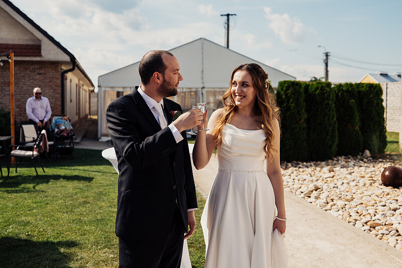 nevesta v bielych šatách, ženích v čiernom obleku, pár s pohármi, slnečný deň na záhrade, svadobná oslava vonku, radostný moment pri toaste. Agropenzión Hubert, Ružindol.