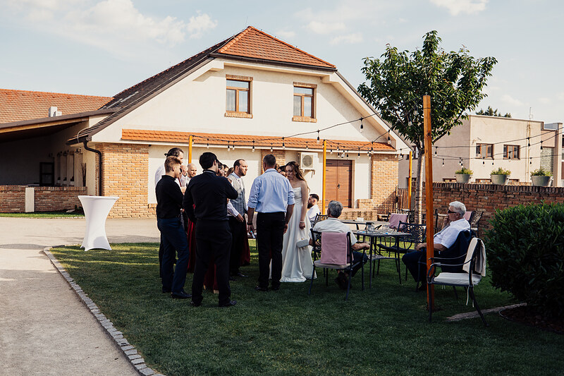 nevesta v bielych šatách, hostia v oblekoch, záhrada s osvetlením, skupinový rozhovor vonku, slnečný deň, stoličky na trávniku. Agropenzión Hubert, Ružindol.