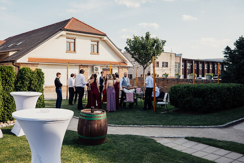 hostia v slávnostnom oblečení, večierok na záhrade, drevený sud s dekoráciou, biely stan s podperami, skupiny ľudí v rozhovore, letný deň s oblačným nebom. Agropenzión Hubert, Ružindol.