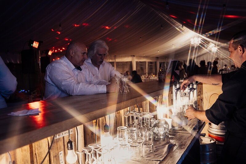 muži pri bare, barman servíruje nápoje, svadobná sála s červeným svetlom, sklenené poháre na pulte, večerná oslava v hale, teplé osvetlenie a atmosféra. Agropenzión Hubert, Ružindol.