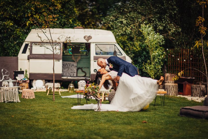 ženích drží nevestu, prvý tanec na trávniku, svadobný pár pred karavanom, slnečný deň v prírode, vintage karavan na pozadí, nevesta v bielych šatách. Hacienda Šišov.