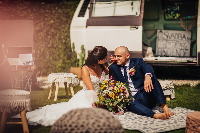 nevesta v bielych šatách, ženích v modrom obleku, pár sedí na trávniku, kvetinová kytica, vintage karavan v pozadí, slnečný deň, intímny moment páru. Hacienda Šišov.