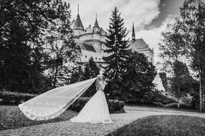nevesta so závojom, pár pred hradom, dlhý svadobný závoj, lesný zámok v pozadí, pár v parku. Meridiana Resort, Bojnice.