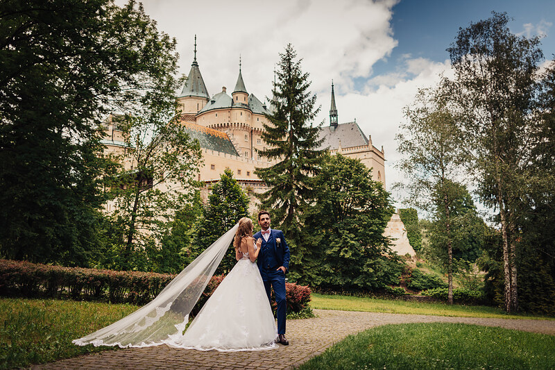 nevesta so závojom, ženích v modrom obleku, zámok v pozadí, pár v parku, dlhá vlečka závoja, slnečný deň s oblakmi. Meridiana Resort, Bojnice.
