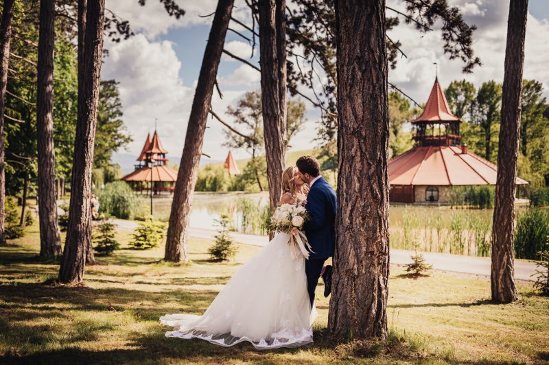 nevesta v bielych šatách, ženích v modrom obleku, bozk medzi stromami, červené strechy v pozadí, slnečný deň s oblakmi, pár pod vysokými borovicami. Meridiana Resort, Bojnice.