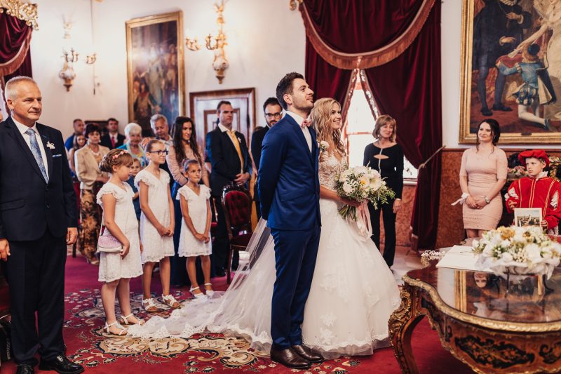 nevesta v bielych šatách, ženích v modrom obleku, slávnostná sála s červeným kobercom, deti v bielych šatách, zlaté zrkadlá a obrazy, hostia v pozadí, kvetinová kytica, slávnostný ceremoniál. Meridiana Resort, Bojnice.