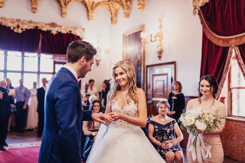 nevesta v čipke, ženích v modrom obleku, výmena prsteňov, slávnostná sála so zlatými dekoráciami, hostia v pozadí, červené závesy a zrkadlá. Meridiana Resort, Bojnice.