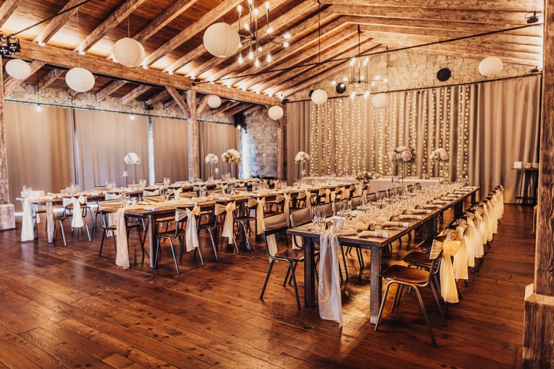 dlhé stoly s hostami, drevené trámy stropu, biele lampióny nad stolmi, svadobná výzdoba s kvetmi, zlaté závesy na pozadí, hostia pri večeri. Meridiana Resort, Bojnice.