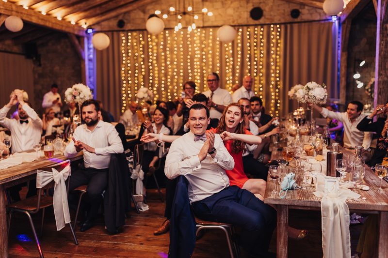 mladý pár tancuje, svadobní hostia sedia, zlaté svetlá na stene, fialové osvetlenie sály, červené šaty nevesty, muž v bielej košeli, veselá atmosféra večera. Meridiana Resort, Bojnice.