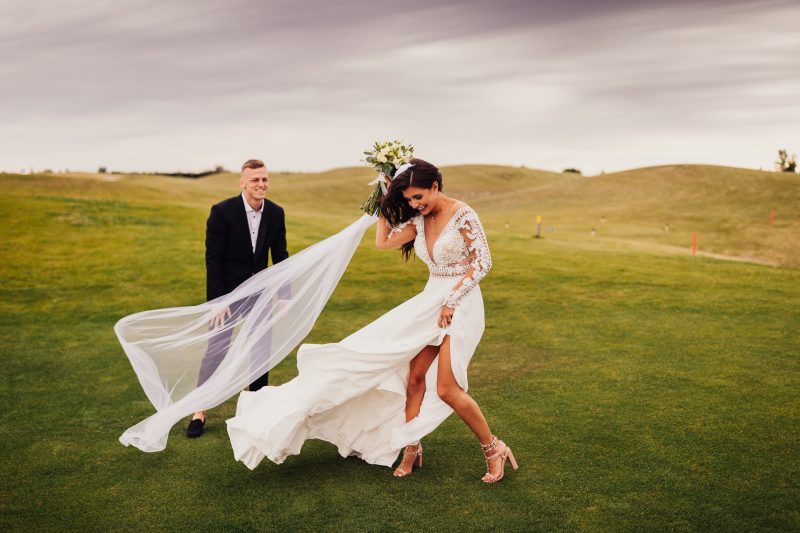 nevesta v bielych šatách, ženích v čiernom obleku, pár na zelenom poli, svadobný závoj vo vetre, slnečný deň na lúke, radosť a smiech. Golf Club Welten, Báč.