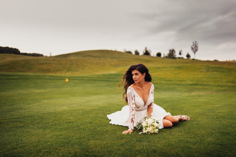 nevesta v bielych šatách, krajková blúzka s gombíkmi, žena sediaca na trávniku, kvetinová kytica, zelené kopce v pozadí, zamračené nebo. Golf Club Welten, Báč.
