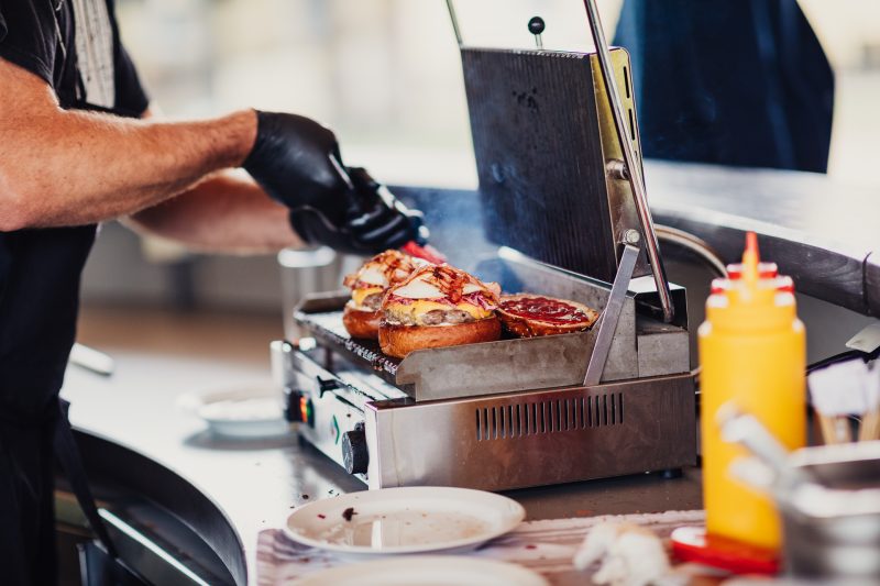 muž griluje mäso, príprava cateringu vonku, oslava za slnečného dňa, kuchár v čiernych rukaviciach, gril s jedlom, žltý nápoj na stole. Golf Club Welten, Báč.