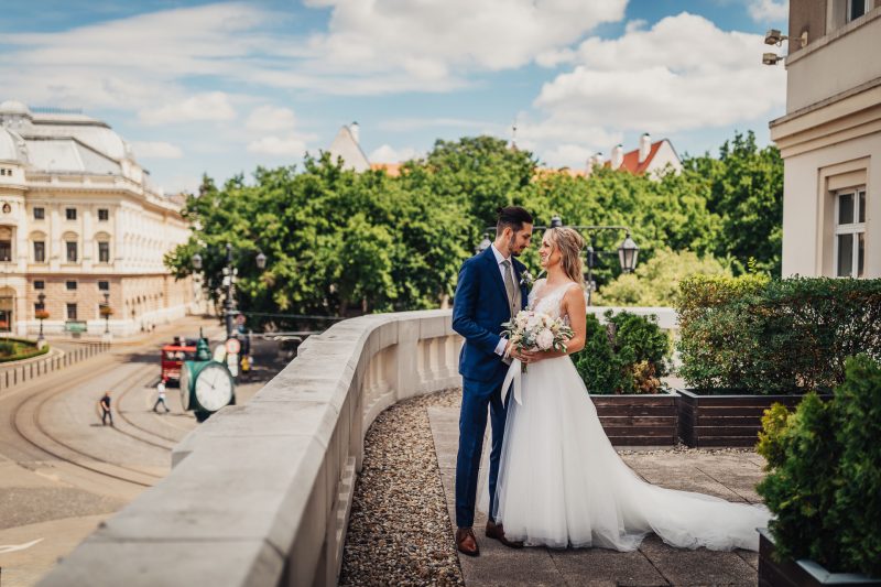 nevesta v bielych šatách, ženích v modrom obleku, pár na moste, mestská architektúra v pozadí, slnečný deň s oblakmi, romantický moment na terase. Karolinenhof Farm, Hainburg.