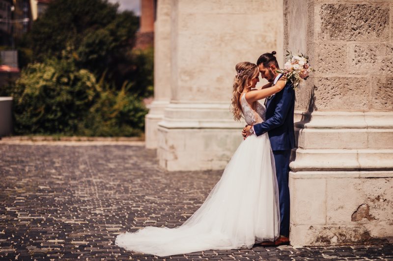 nevesta v bielych šatách, ženích v modrom obleku, pár pri kamennej stene, dlhý svadobný závoj, slnečný deň, romantický moment pri stene. Karolinenhof Farm, Hainburg.