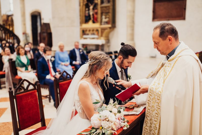 nevesta v závoji pri oltári, kňaz čítajúci z knihy, svadobný obrad v kostole, ženích vedľa nevesty, červené stoličky v chráme, biele kvety na oltári. Karolinenhof Farm, Hainburg.