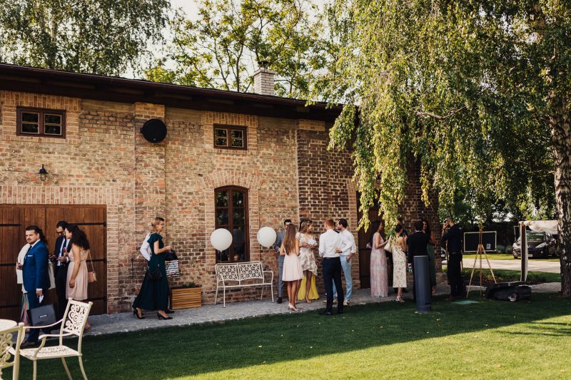 hostia v slnečnom svetle, tehlový dom s brečtanom, svadobní hostia v elegantných šatách, biele balóny pri vchode, skupiny ľudí v rozhovore, trávnik pri starom dome. Karolinenhof Farm, Hainburg.