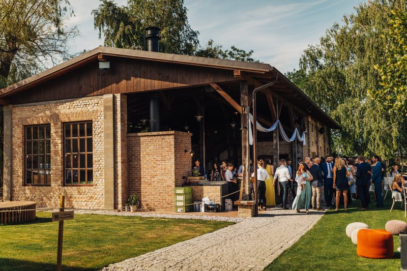 hostia v slnečnom svetle, svadobný ceremoniál vonku, tehlový dom s dekoráciami, ľudia v elegantných šatách, slávnostná atmosféra na trávniku, modrá výzdoba pod strechou. Karolinenhof Farm, Hainburg.