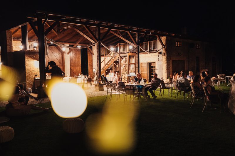 nočná svadba na dvore, hostia sedia pri stoloch, teplé svetlo pergoly, osvetlené guľové lampy, večerná slávnosť vonku, tehlový dom s osvetlením. Karolinenhof Farm, Hainburg.
