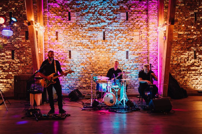 hudobná skupina na pódiu, muž spieva s gitarou, bubeník pri hre, tehlová stena s osvetlením, ružové a modré svetlá, interiér s drevenou podlahou. Karolinenhof Farm, Hainburg.