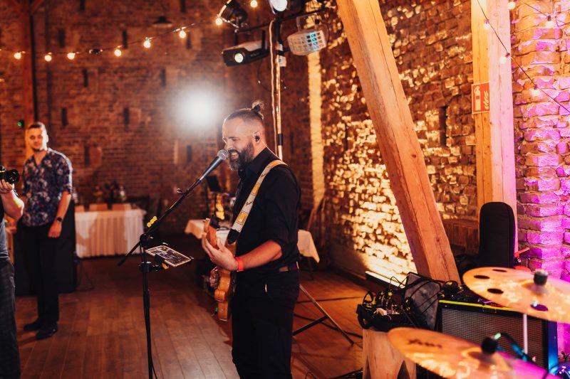 gitarista spieva, tehlová stena s ružovým svetlom, večerná svadba v pivnici, muž s bradou pri mikrofóne, teplé osvetlenie a girlandy, hostia počúvajúci hudbu. Karolinenhof Farm, Hainburg.
