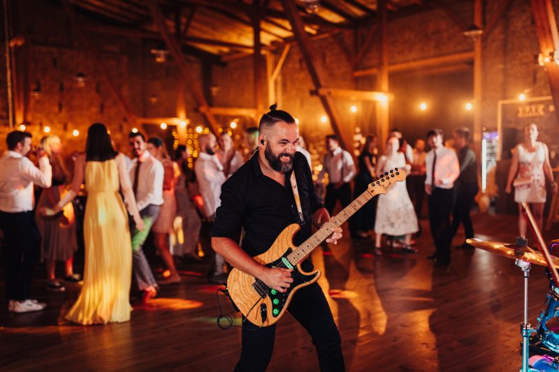 gitarista hrá na svadbe, hostia tancujú v noci, teplé osvetlenie v areáli, usmiaty muž s gitarou, večerná svadba s hudbou, zlaté svetlá nad tanečným parketom. Karolinenhof Farm, Hainburg.