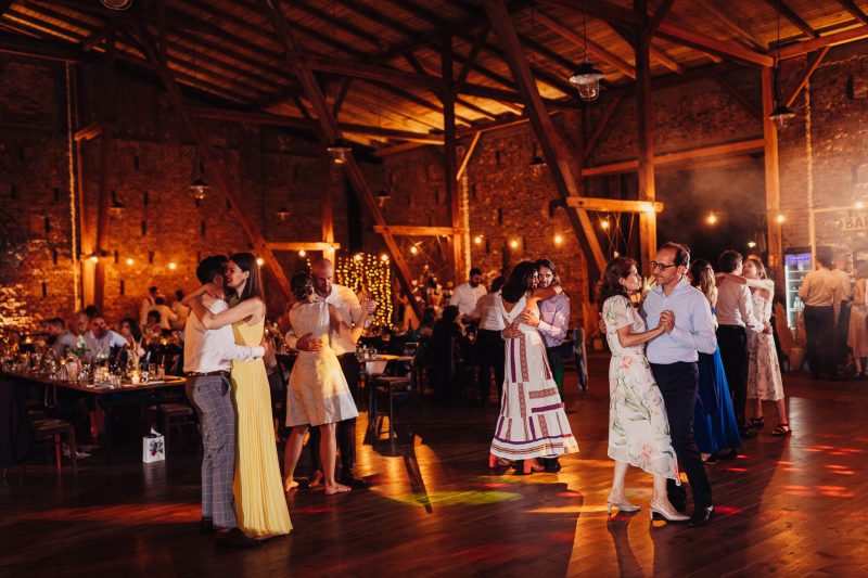 páry tancujú na parkete, večerná svadba v stodole, teplé osvetlenie žiarovkami, hostia tancujú spolu, kamenná budova s drevom, zlaté svetlo večera. Karolinenhof Farm, Hainburg.