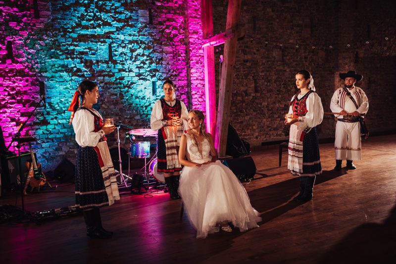 nevesta v bielych šatách, tradičný ľudový odev, kamenná stena s osvetlením, ružové a tyrkysové svetlá, svadobný ceremoniál v interiéri, spev a hudba na pódiu. Karolinenhof Farm, Hainburg.