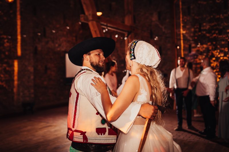 mladomanželia v ľudovom kroji, prvý tanec v noci, biele a červené tradičné oblečenie, teplé svetlo lampiónov, svadba pod hviezdami, pár tancujúci vonku. Karolinenhof Farm, Hainburg.