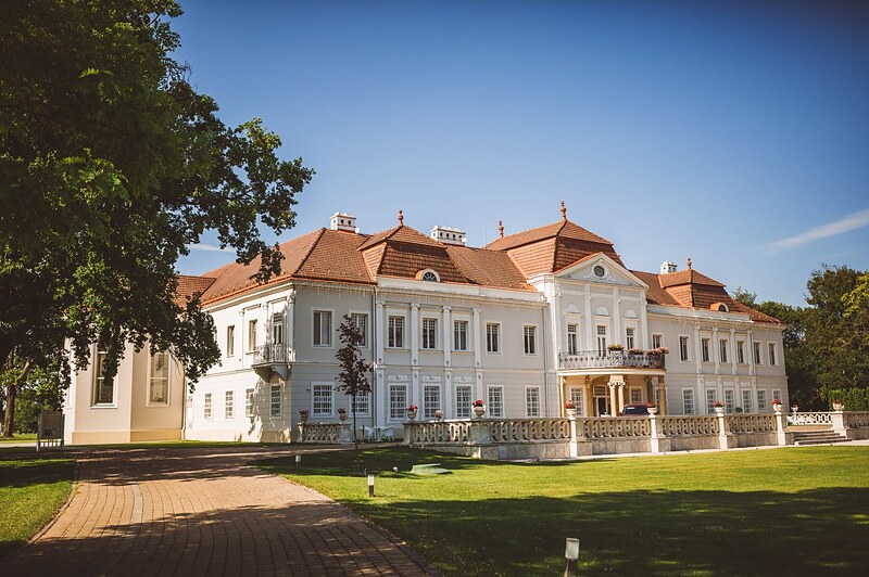 biely barokový kaštieľ s červenou mansardovou strechou, trávnik a dláždená cesta pred vchodom. Art Hotel Kaštieľ Tomášov.