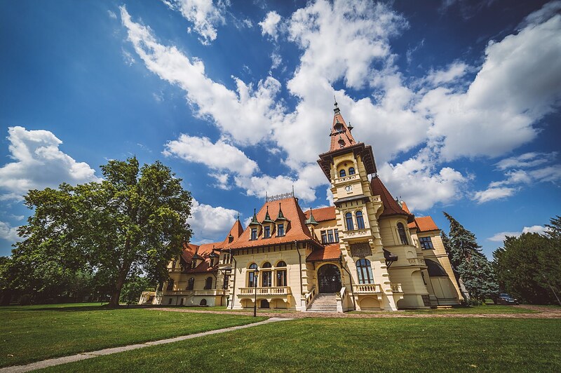 historická budova s vežičkou a červenou strechou v parku, biele oblaky na modrej oblohe. Art Hotel Kaštieľ Tomášov.
