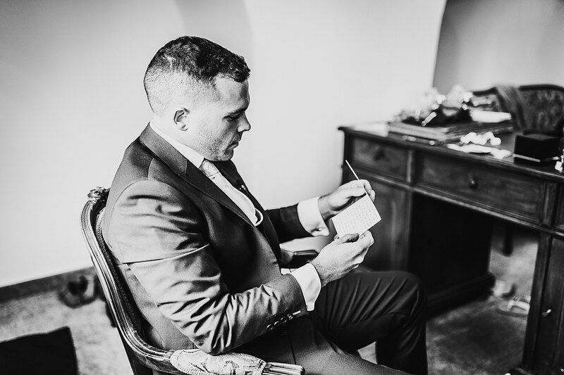 čítajúci muž v obleku, hostia sediaci pri stole, formálny interiér, zamyslený výraz, vintage atmosféra. Kaštieľ Villa Bork, Borčice.