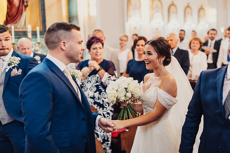 nevesta s kvetmi v kostole, ženích v modrom obleku, pár si drží ruky, svedkovia v pozadí, sviečky v chráme, radostný výraz nevesty. Kaštieľ Villa Bork, Borčice.