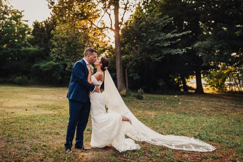 nevesta a ženích sa bozkávajú, pár v prírode pri západe slnka, svadobné šaty s vlečkou, zlaté svetlo medzi stromami, romantický moment v parku, večerný bozk v prírode. Kaštieľ Villa Bork, Borčice.