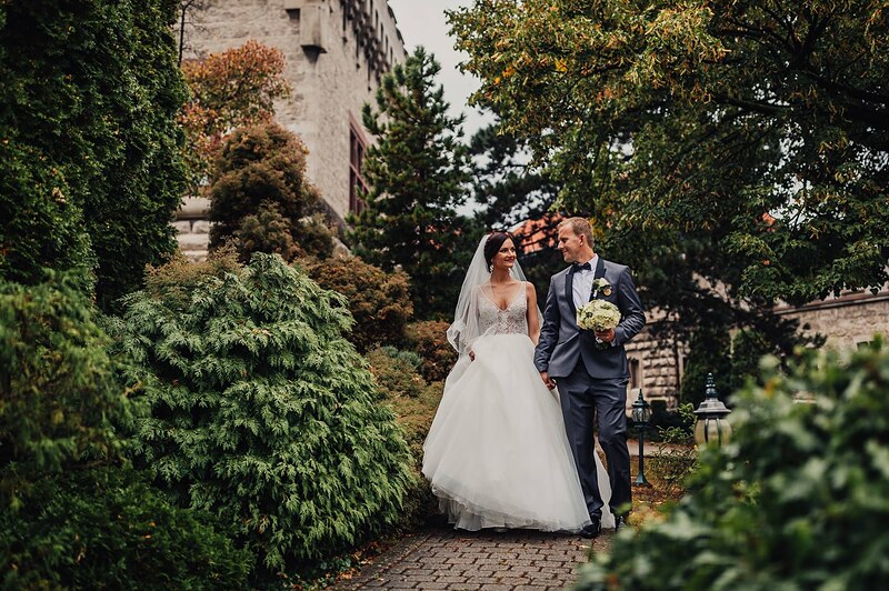 pár kráča po chodníku medzi zeleňou, ženích v tmavomodrom obleku, kamenná stavba v pozadí. Hotel Jeleň, Hlohovec.