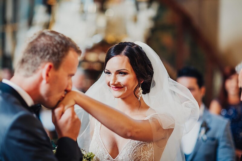 ženích bozkáva neveste ruku počas obradu, hostia okolo, závoj a čipkované šaty. Hotel Jeleň, Hlohovec.