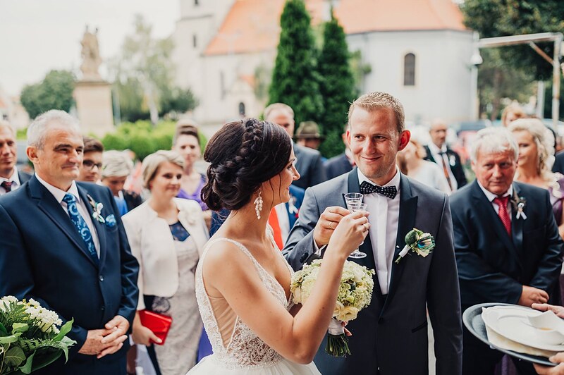 prípitok pred kostolom, novomanželia s pohármi, hostia okolo, veža kostola v pozadí. Hotel Jeleň, Hlohovec.