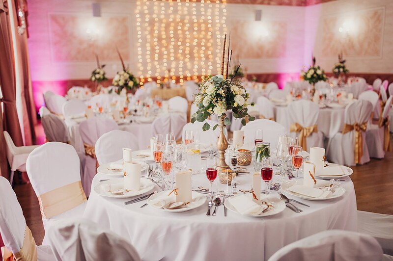 prestretá svadobná sála, okrúhle stoly so zlatými stuhami, vysoké kvetinové aranžmány a svetelný záves. Hotel Jeleň, Hlohovec.