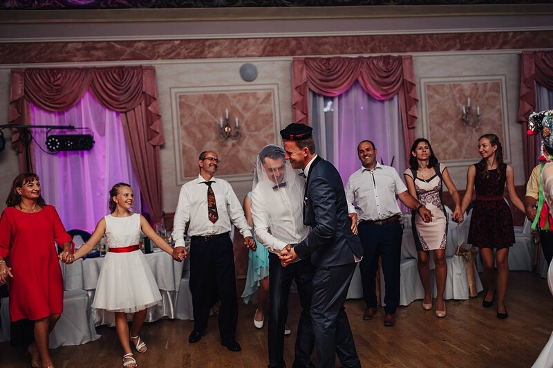 hostia sa držia za ruky v kruhu okolo novomanželov, ženích v klobúku, veselá sála. Hotel Jeleň, Hlohovec.