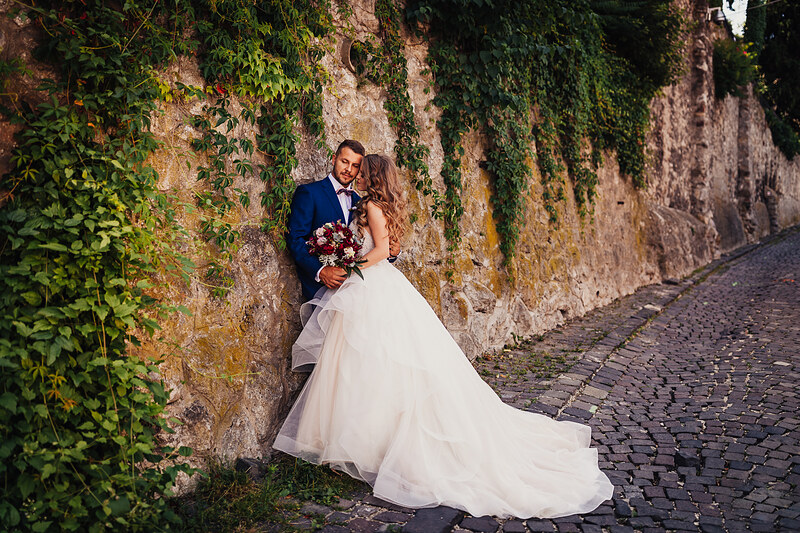 nevesta v bielych šatách, ženích v modrom obleku, pár pri starom múre, popínavé rastliny na stene, kamenná ulica, teplé denné svetlo. Kaštieľ Klobušice.