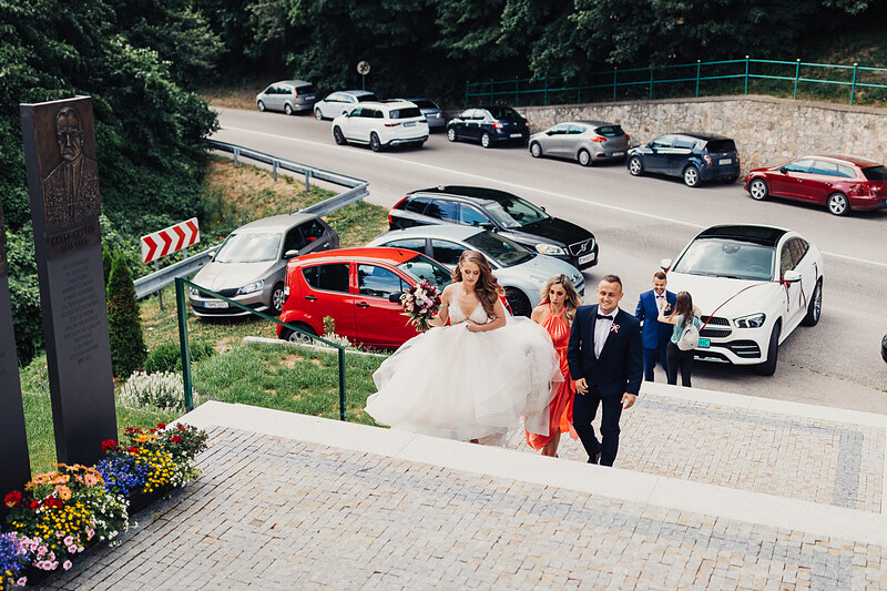 nevesta v bielych šatách, ženích v modrom obleku, svadobný sprievod na parkovisku, červené auto medzi hosťami, slnečný deň na asfaltovej ploche, formálna svadobná ceremónia. Kaštieľ Klobušice.