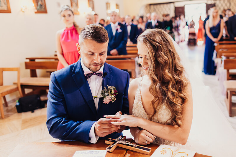 ženích a nevesta si vymieňajú prstene, kostol počas obradu, nevesta v čipkovaných šatách, ženích v modrom obleku, slávnostný okamih v chráme, hostia v pozadí. Kaštieľ Klobušice.
