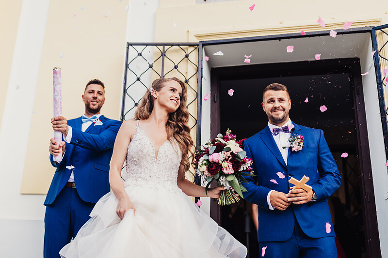 nevesta v bielych šatách, ženích v modrom obleku, papierové konfety padajúce, pár pred kostolom, slnečné svetlo na tvári, radostná atmosféra svadby. Kaštieľ Klobušice.
