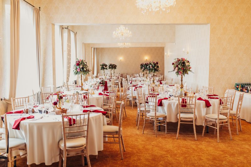 svadobná sála s tabuľkami, okrúhle stoly s bielymi obrusmi, červené dekorácie na stoloch, kvetinové aranžmány na stojane, krištáľové lustre nad sálou, zlaté stoličky pri tabuľkách. Kaštieľ Klobušice.