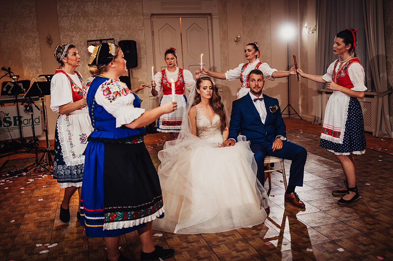 nevesta v bielych šatách, ženích v modrom obleku, tradičný ľudový tanec, kultúrny program na svadbe, sála s teplým osvetlením, sediaci pár počas predstavenia. Kaštieľ Klobušice.