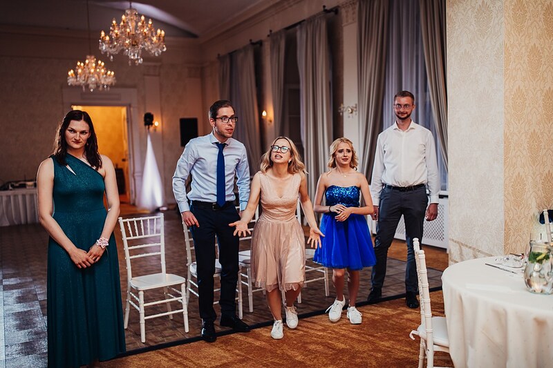hostia sa smejú počas svadobnej hry so stoličkami, luster a závesy v sále. Kaštieľ Klobušice.