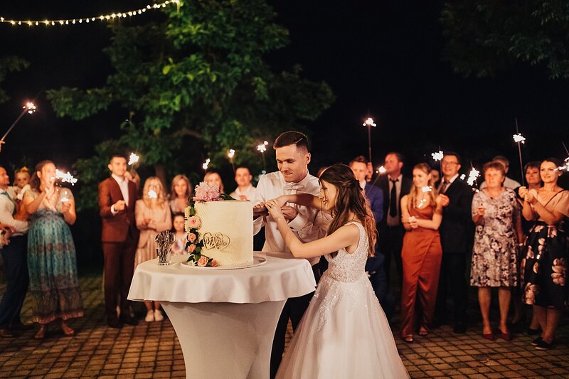 krájanie torty vonku, hostia okolo držia prskavky, svetelné reťaze v korunách stromov. Kaštieľ Klobušice.