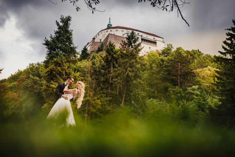 ženích drží nevestu, nevesta v bielych šatách, hrad v pozadí, les okolo páru, oblačné nebo, romantická chvíľa v prírode. Hotel Zlatý Kľúčik, Nitra.